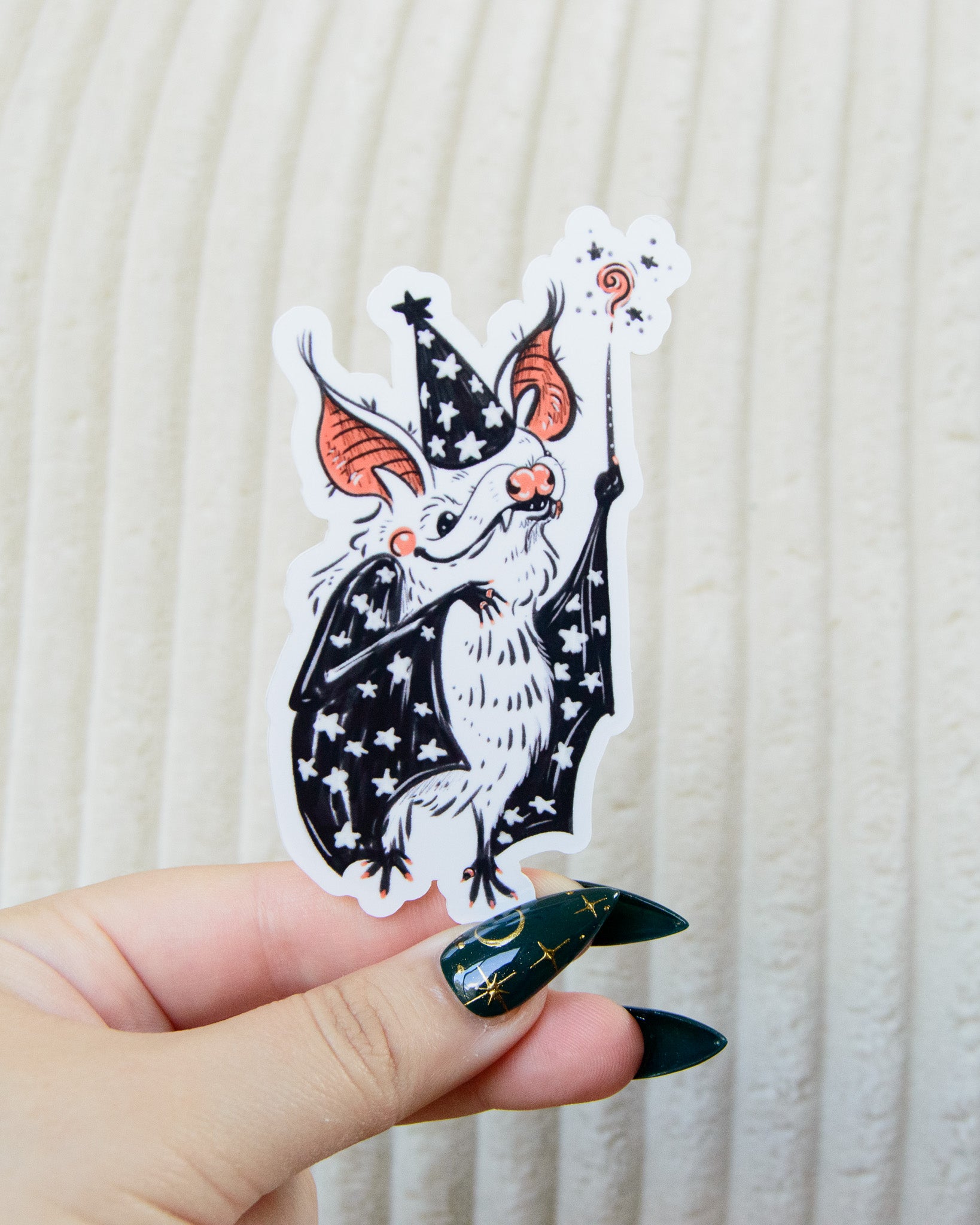 Hand holding a sticker of a cartoon bat character with a star witch wizard hat and a magic wand