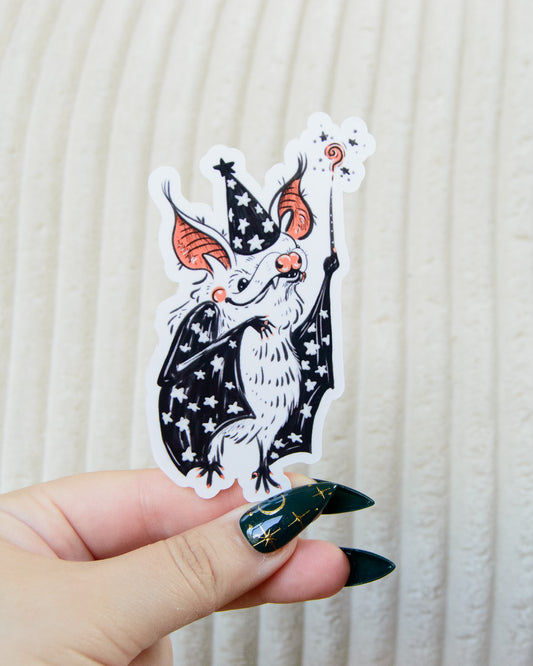 Hand holding a sticker of a cartoon bat character with a star witch wizard hat and a magic wand